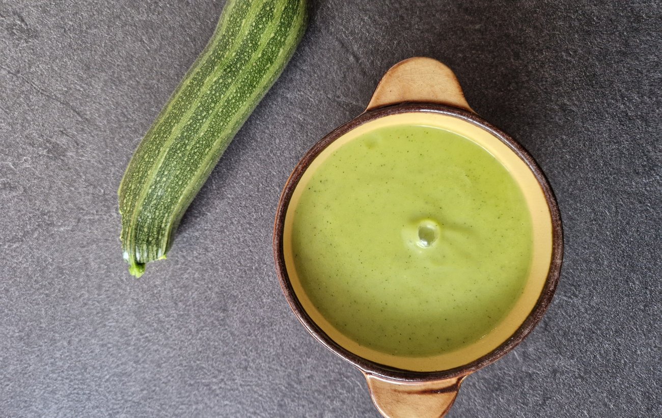 Velouté de courgette à la vache qui rit servi dans une cocotte en terre, à côté d'une courgette entière